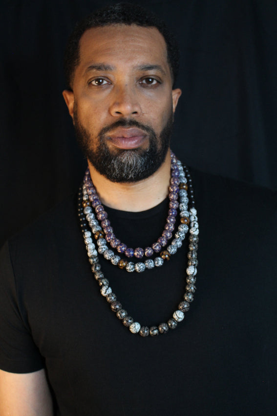 A man with a beard wearing a black t-shirt and multiple layered beaded necklaces, photographed against a black background.